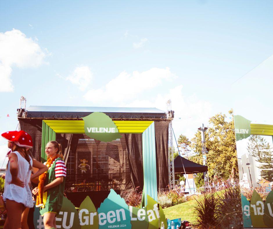 Outdoor event stage with "EU Green Leaf" and "Velenje 2024" banners. Several individuals are in the foreground, one wearing a mushroom costume. Clear blue sky and trees in the background.