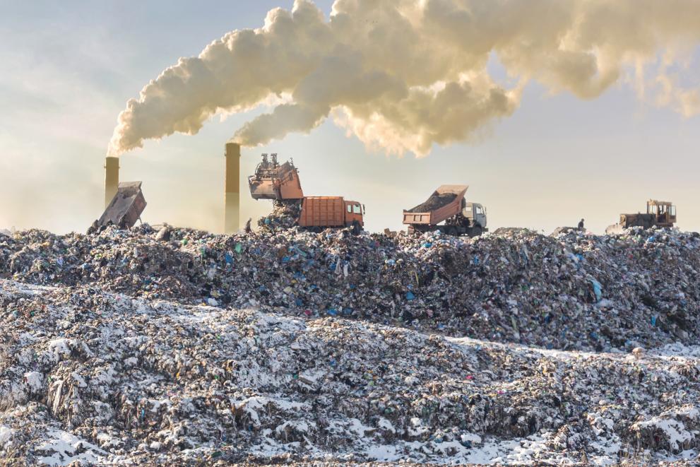 Dump trucks unloading garbage over vast landfill with smoking industrial stacks in the background